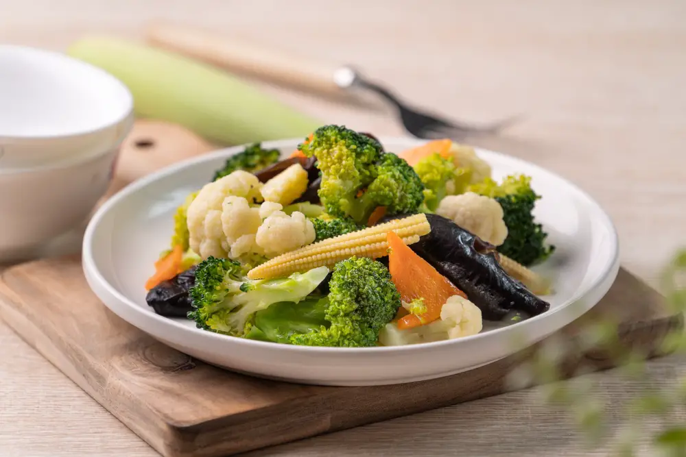 A plate of boiled vegetables, including corn, broccoli, and cauliflower, sitting on a piece of wood on a table.
