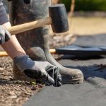 A person kneeling and installing landscape fabric with garden stakes and a mallet.