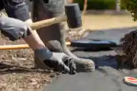 A person kneeling and installing landscape fabric with garden stakes and a mallet.