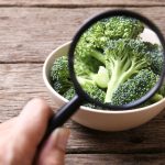 Someone inspecting broccoli in a bowl on a table with a magnifying glass.