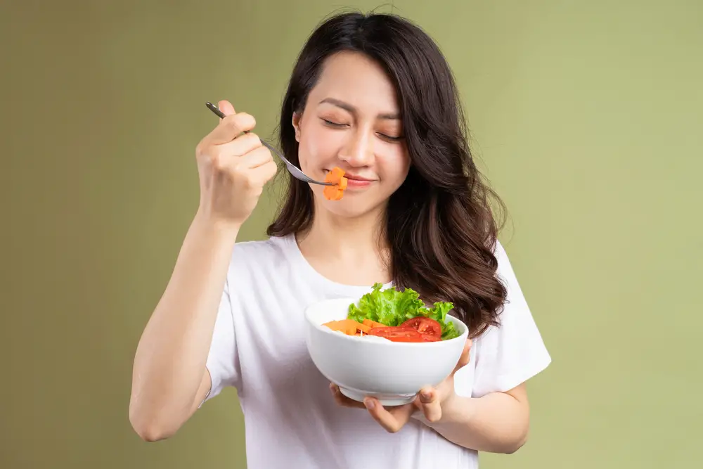 A happy woman about to eat a carrot from a bowl of carrots, lettuce, and tomatoes.