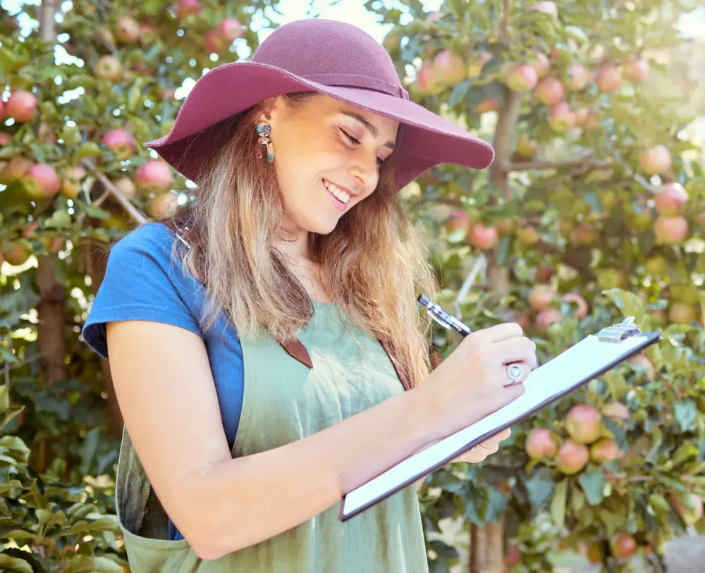 A happy woman writing on a clipboard on a sunny day in an apple orchard.