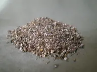 A mound of pale brown and silver vermiculite flakes on a gray surface, showing the layered texture of the ingredient.